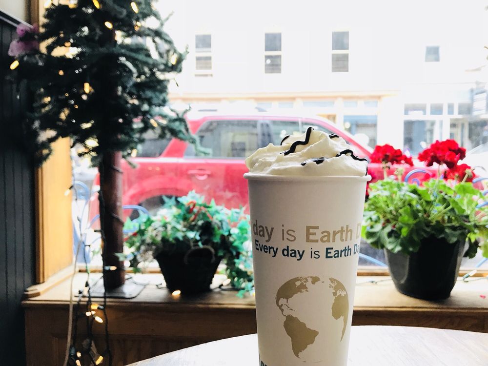 Coffee cup with whipped cream in front of window seat with lots of flowers looking out on street